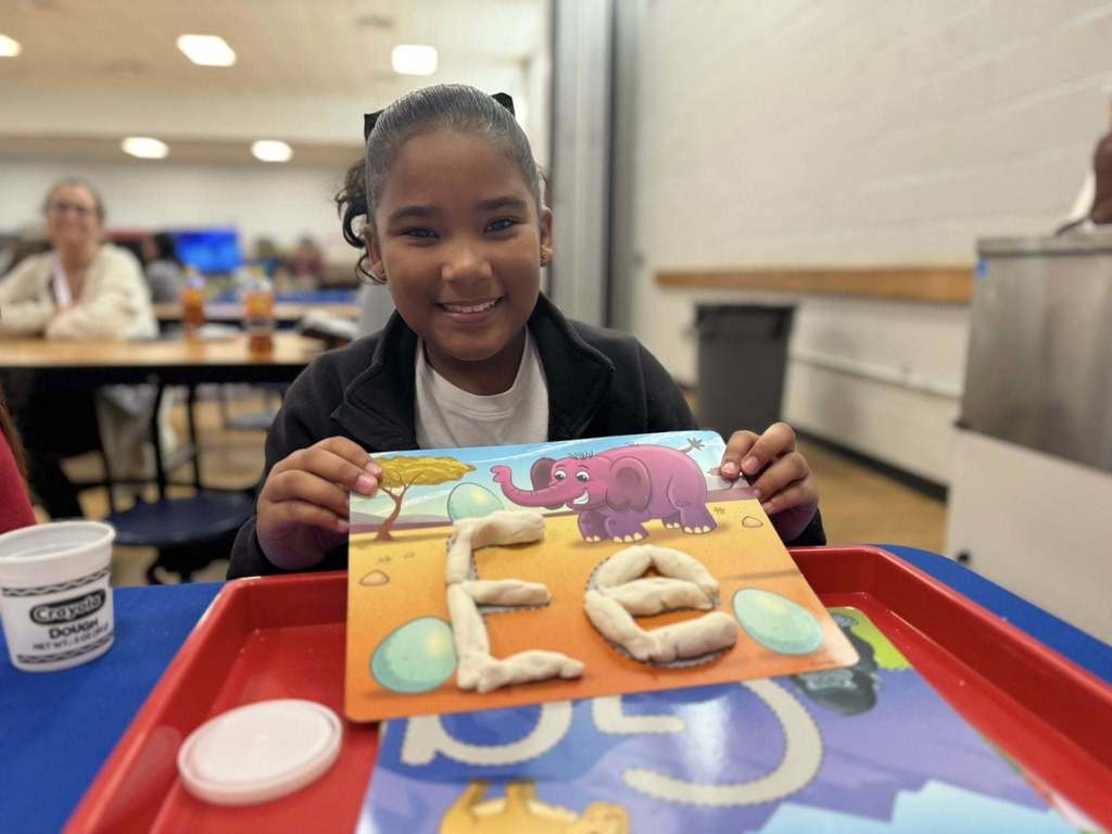 A smiling girl is holding up an orange mat with a clay elephant and the letters "E" and "L" made from dough in a school cafeteria. A person can be seen in the background.