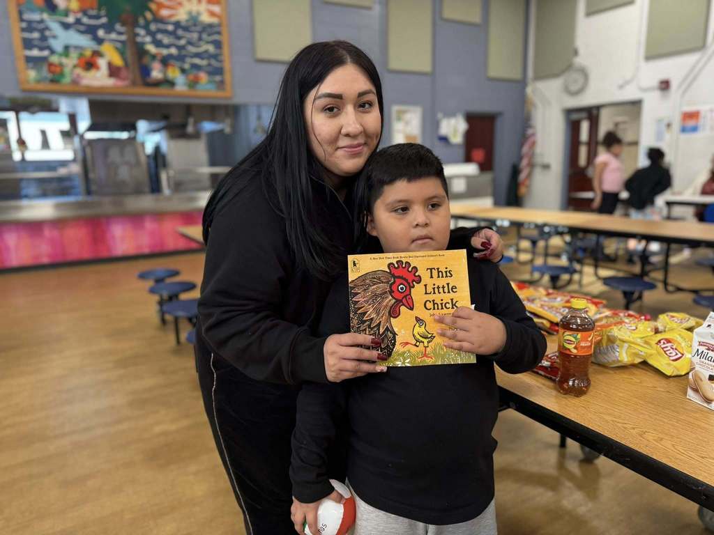 An adult and a young boy are standing in a school cafeteria holding a book titled "This Little Chick." Other children can be seen in the background.