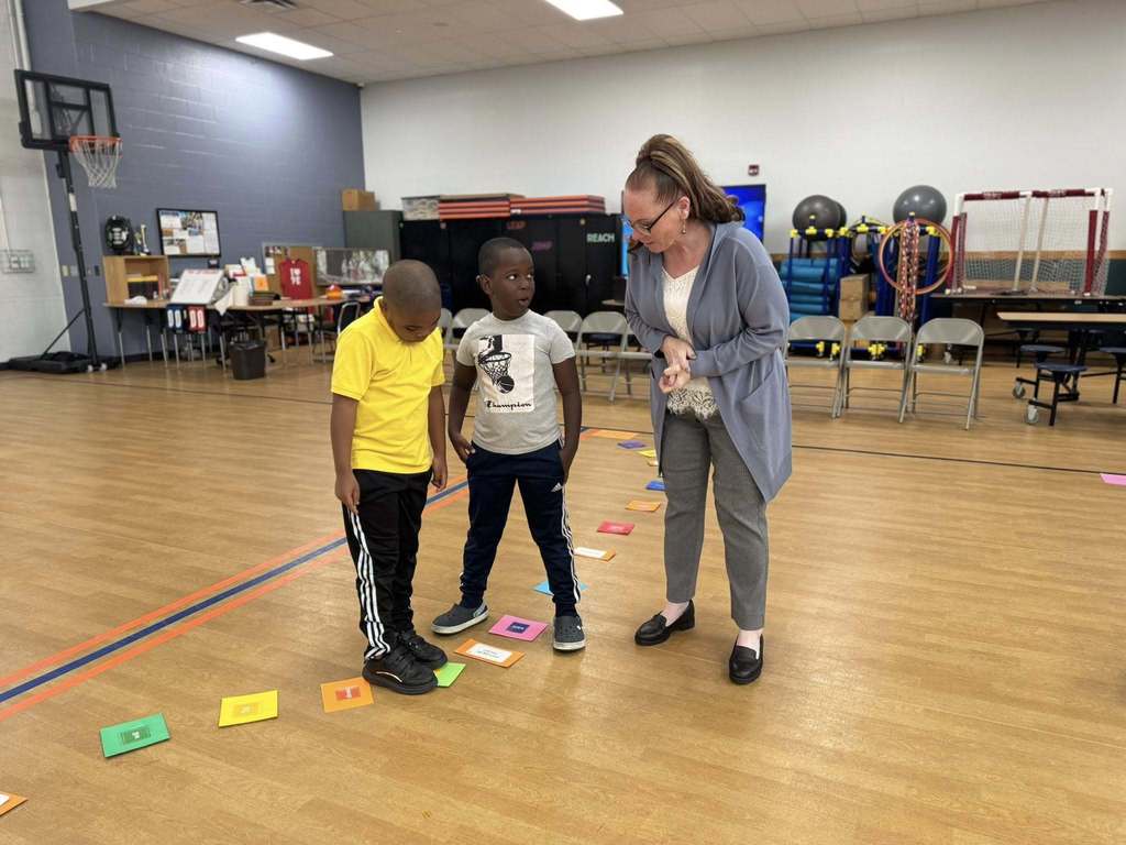 A district staff member is guiding two young boys in an activity in a school gym with colorful squares.