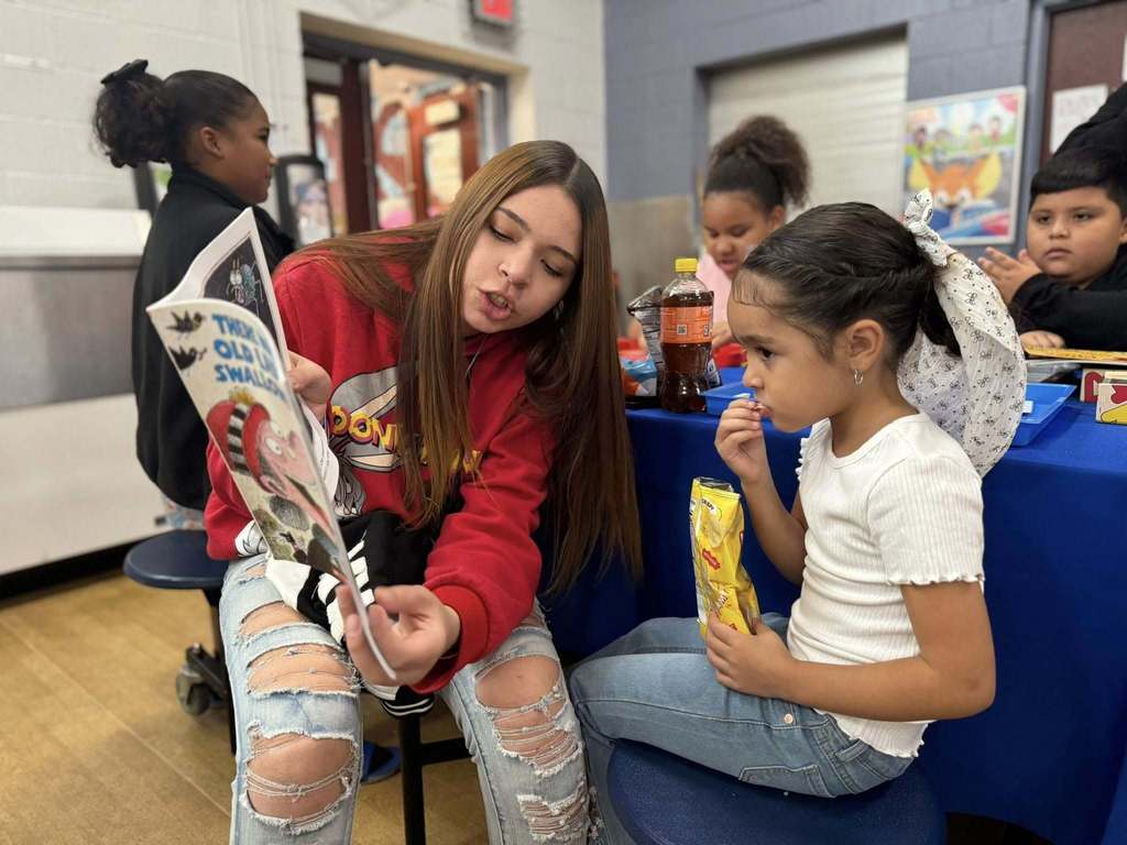 A young woman is sitting down reading a book to a young girl who is also sitting down at a table eating a bag of chips in a school cafeteria. Other children can be seen in the background.