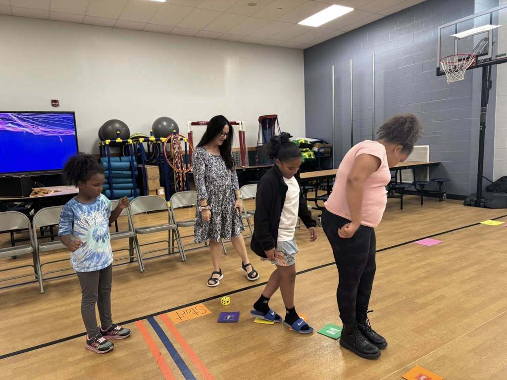Three young girls and an adult are engaging in a group activity in a school gym using exercise equipment, chairs, and a basketball hoop.