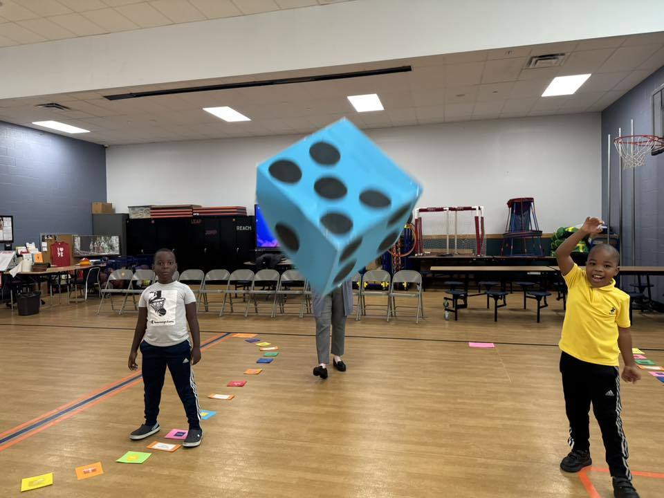 A district staff member and two young boys are playing in a school gym. One of the boys is throwing a large blue die. Colorful cards can be seen nearby on the floor.