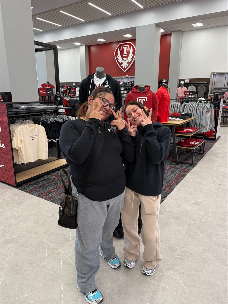Two young women are holding up peace signs in a university apparel store. The background features red and gray hoodies with a university logo.