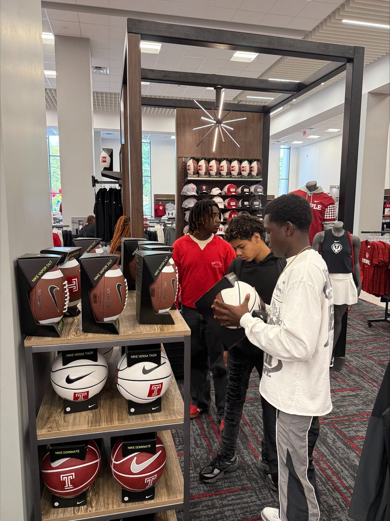 A group of three young men are looking at sports balls in a store, while another young man is looking off into the distance. The shelves are displaying footballs and basketballs with other sports apparel visible.