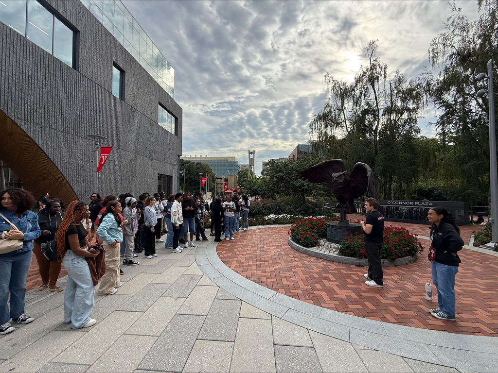 A group of students are standing attentively near a building in O’Connor Plaza, facing a speaker beside a large eagle statue.