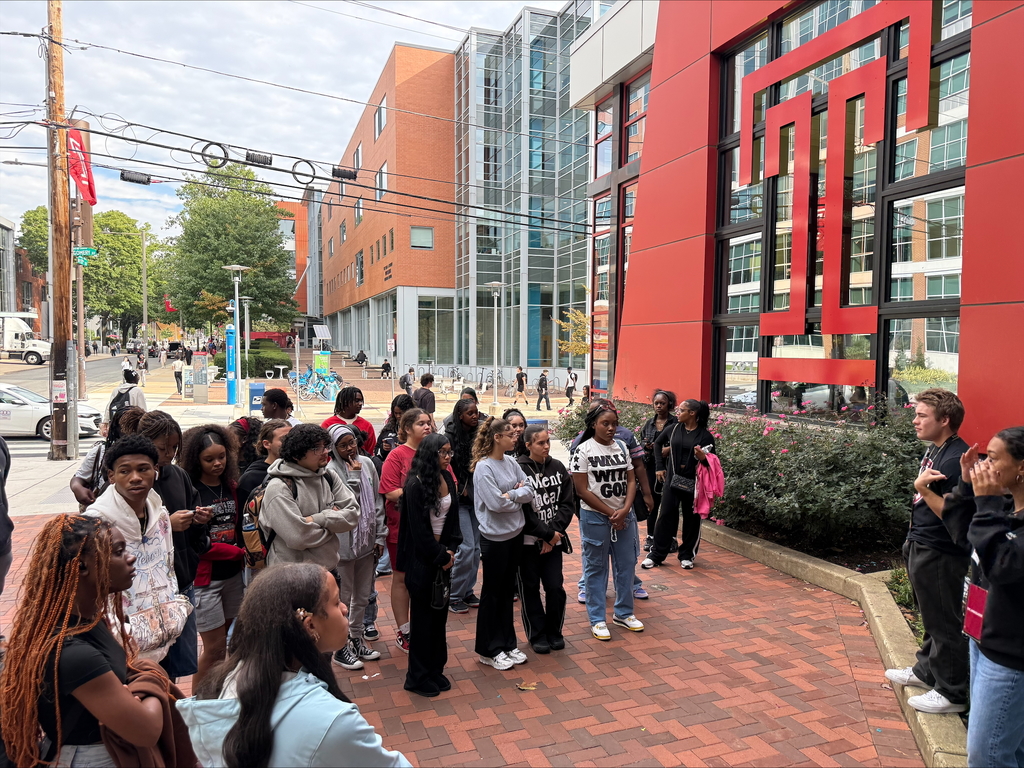 A diverse group of students are standing outside, attentively listening to a speaker at a university campus. The red and white logo of Temple University is visible on a nearby building.