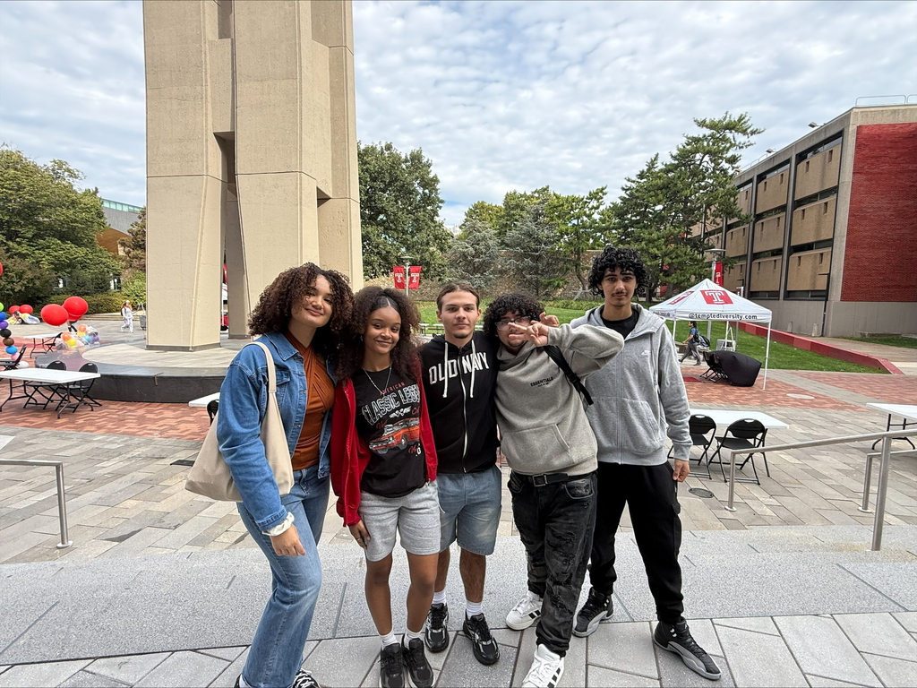 A group of two young women and three young men standing in front of a tall monument on a college campus. Balloons and tents can be seen in the background.