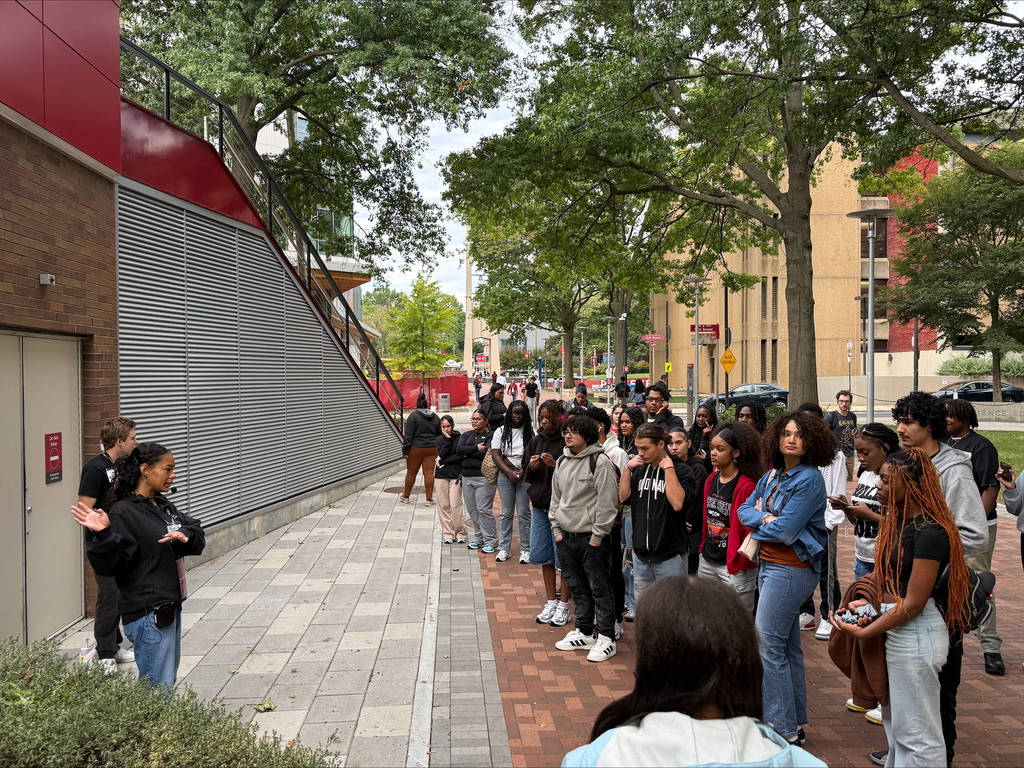 A group of diverse students are standing attentively on a brick path under leafy trees. A person on the left is speaking with gestures.