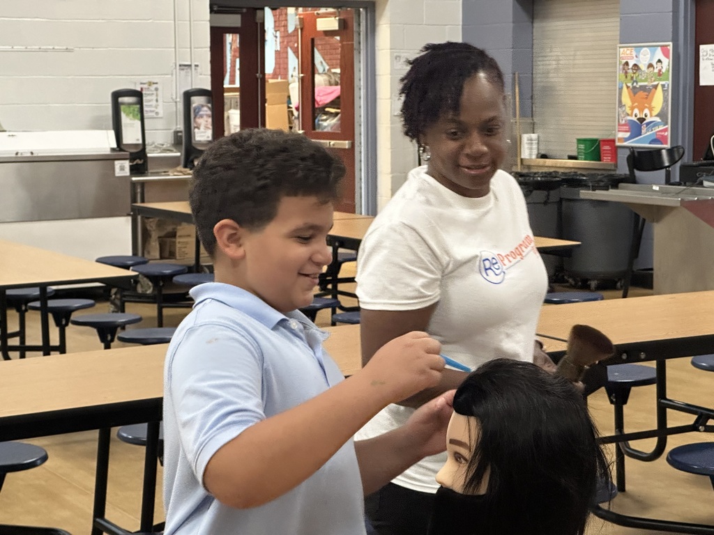 A student is working on a mannequin's head at a table with an instructor observing nearby.