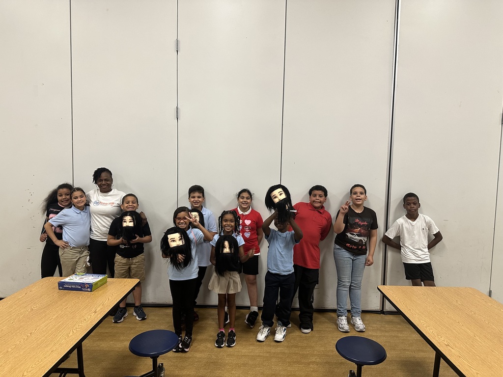 A group of students are standing in a row indoors, some holding masks, in front of a plain wall with tables in the foreground.
