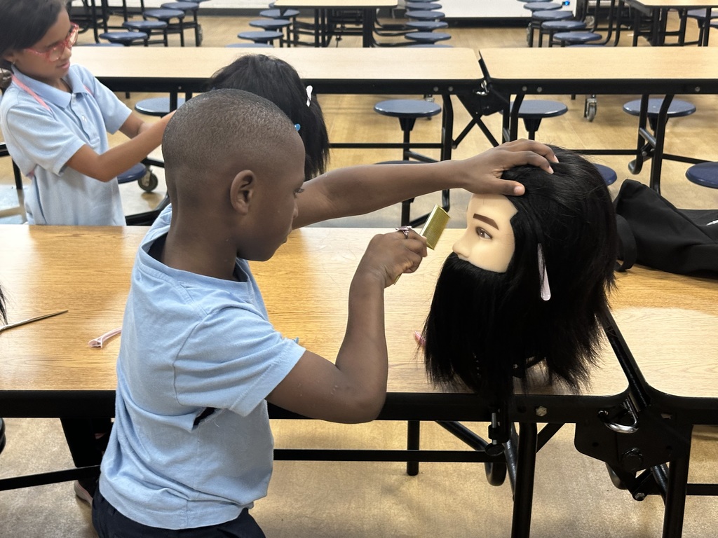 A student is using a comb on a mannequin's head at a table, with another student working on mannequins in the background.