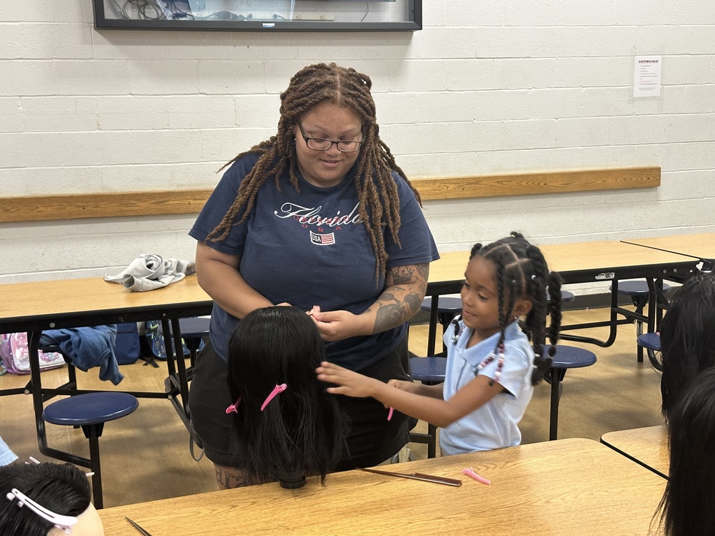 A student is working on a mannequin's head at a table with an instructor beside her helping.