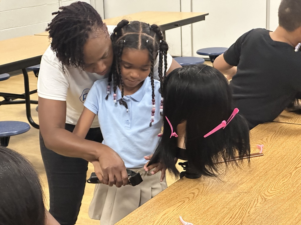 A student is working on a mannequin's head at a table, with an instructor standing over her helping. Another student can be seen in the background.