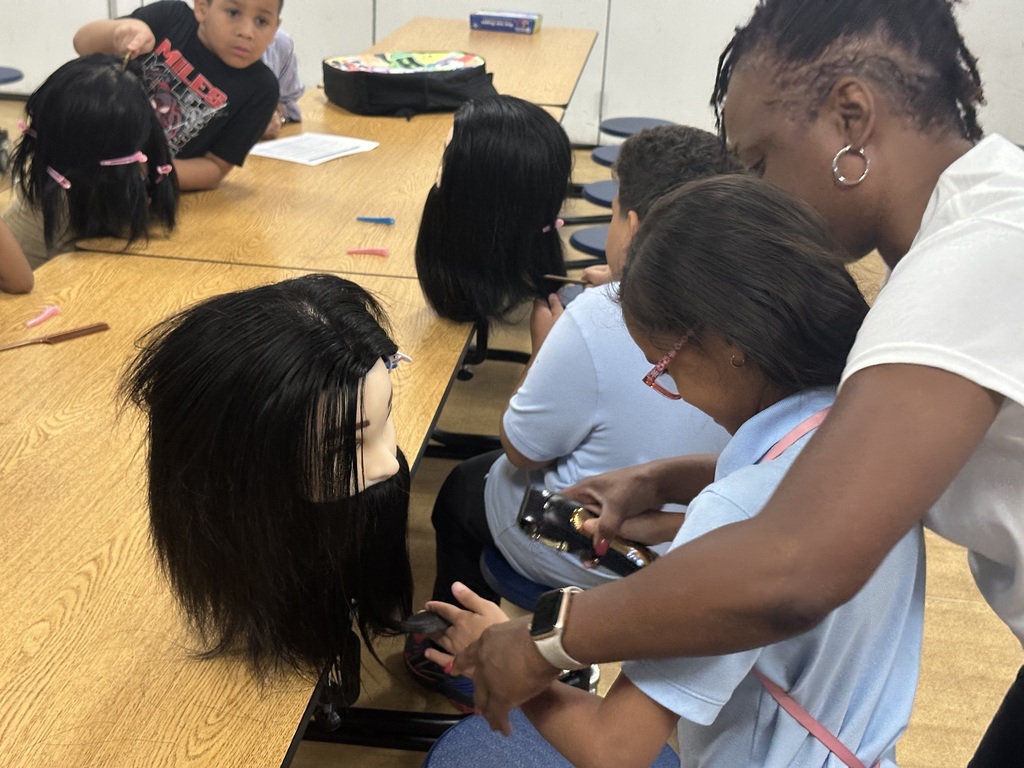 A student is working on a mannequin's head at a table, with an instructor standing over her helping. Other students can be seen working on mannequins in the background.