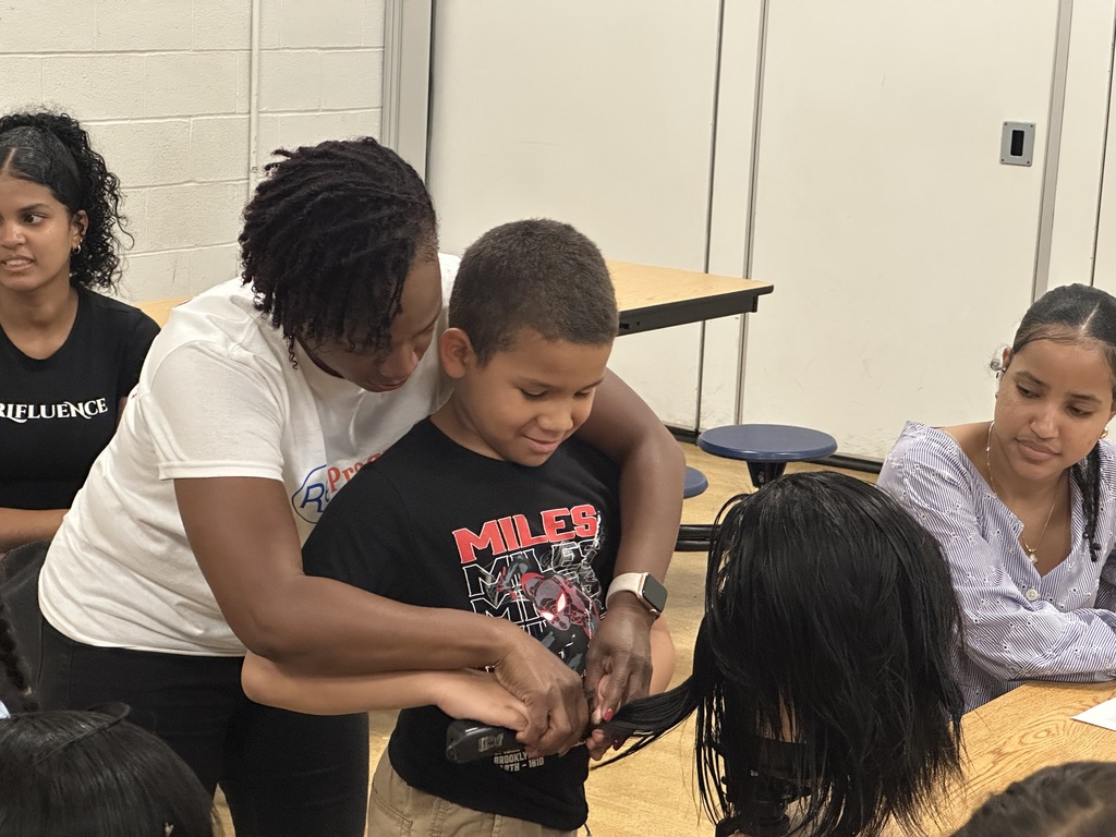 A student is working on a mannequin's head at a table, with an instructor standing over him helping. Two young women can be seen in the background.