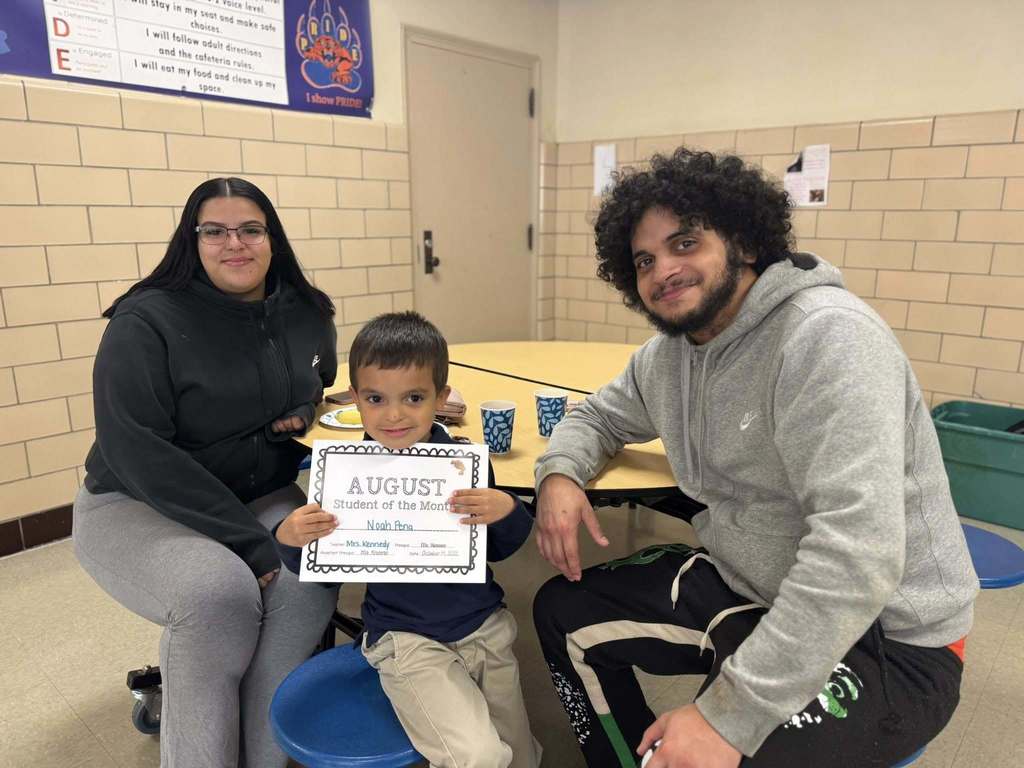 A male student is holding an "August Student of the Month" certificate, accompanied by two adults in a school cafeteria.