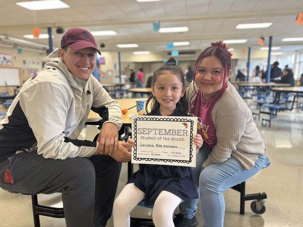 A female student is holding a "September Student of the Month" certificate in a school cafeteria, accompanied by two adults.