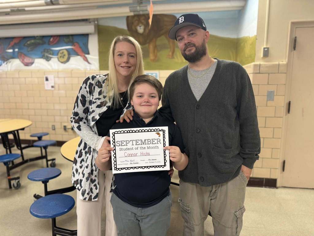 A male student is holding a " September Student of the Month" certificate, standing between two adults in a school cafeteria.