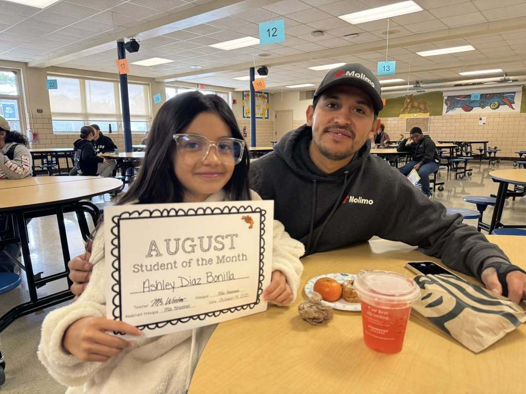 A female student is sitting at a table holding an "August Student of the Month" certificate in a school cafeteria, accompanied by an adult in a school cafeteria. There is a plate of food, a drink and other people nearby.
