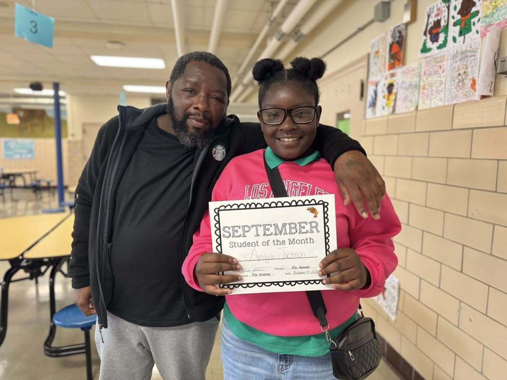 A female student is holding a "September Student of the Month" certificate, accompanied by an adult in a school cafeteria. Artwork can be seen hanging nearby on a wall.