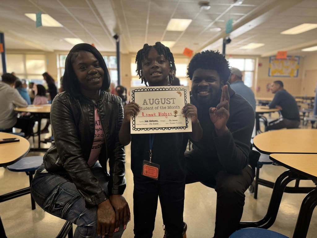 A male student is holding an "August Student of the Month" certificate in a school cafeteria, accompanied by two adults.