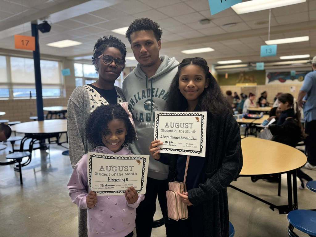 Two female students are holding "August Student of the Month" certificates in a school cafeteria, accompanied by two adults.