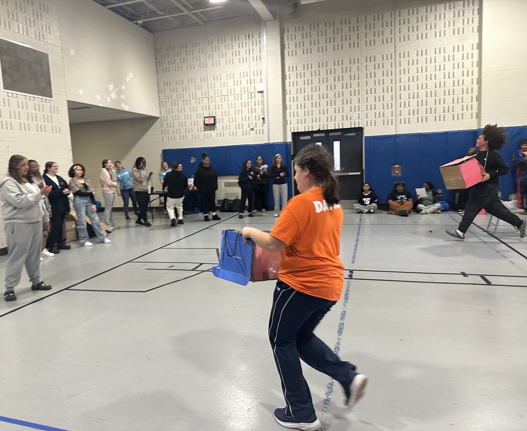 A lively relay race in a school gym featuring student participants carrying colorful bags. Onlookers are cheering from the sidelines.