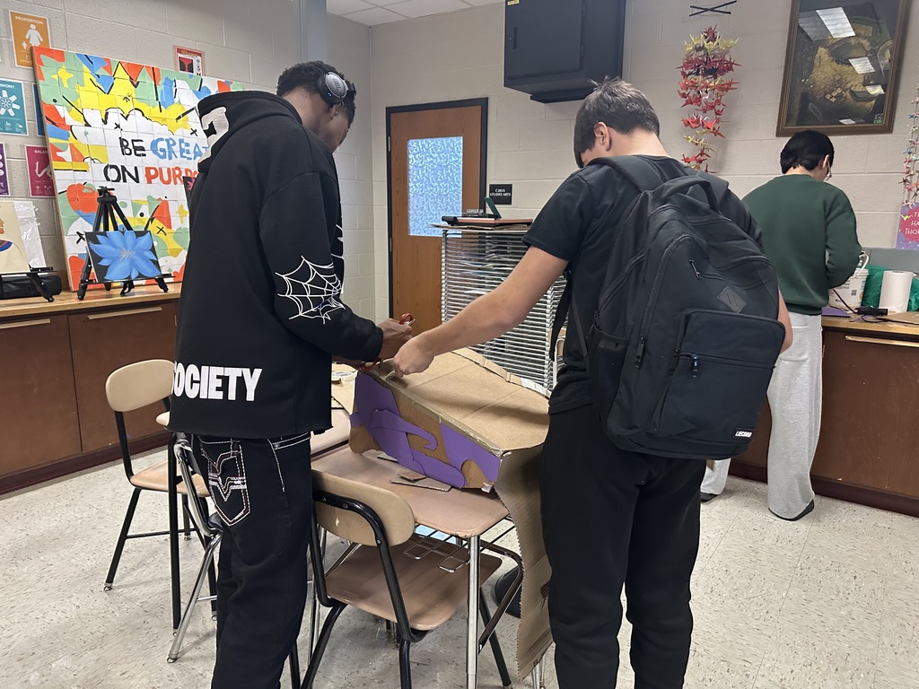 Two students are working together on a cardboard project in a classroom. There is a motivational poster and another student in the background.