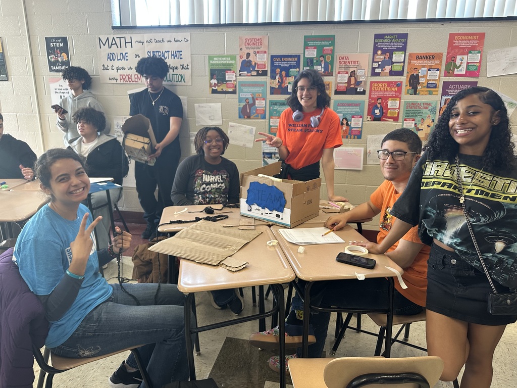 A group of students are gathered around desks in a classroom, engaging in a creative project with cardboard and crafts.