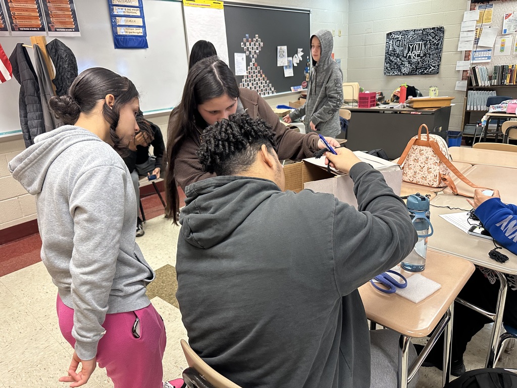 A group of students are collaborating in a classroom, focusing on a project. The group is gathered around a table with scissors, a box, and water bottles.