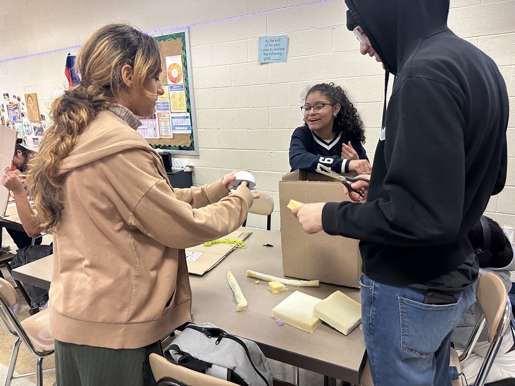 Three students are engaging in a collaborative project in a classroom. They are surrounded by cardboard and tools, working together.