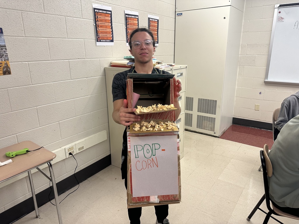 A student standing in a classroom is holding a homemade cardboard popcorn machine filled with popcorn. The machine has a hand-drawn sign that reads "POP-CORN."
