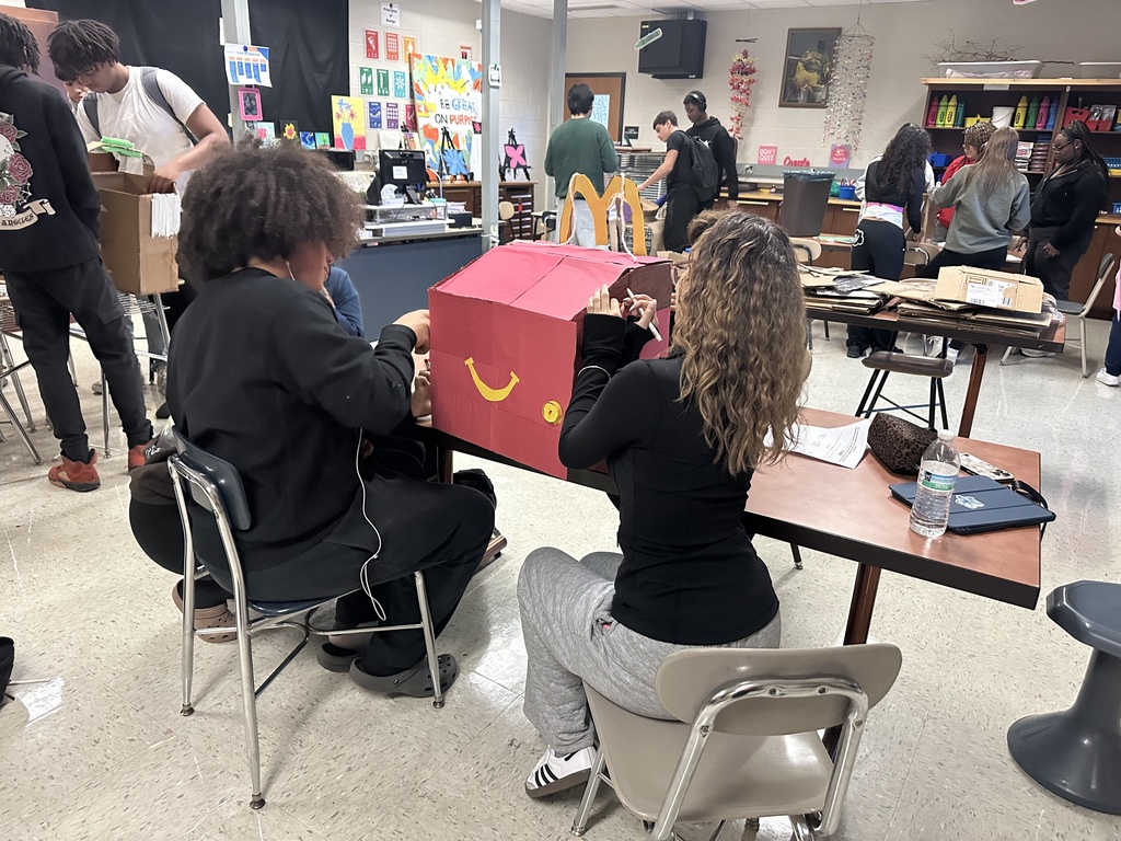 A group of students in a classroom are working collaboratively on a large red project shaped like a fast-food box. Other students can be seen in the background.