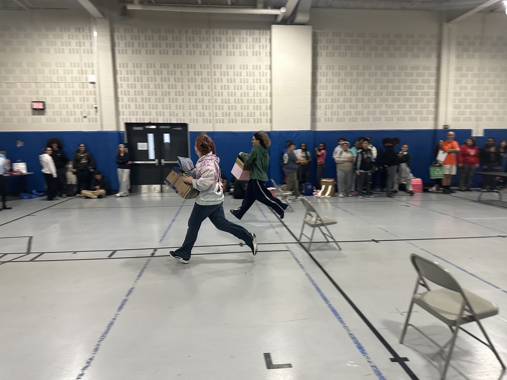 Two students are running inside of a school gym, each of them holding boxes. A line of people are watching in the background.