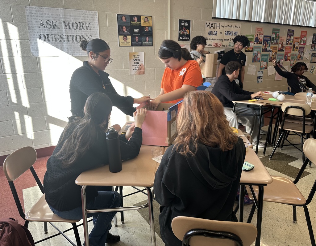 A group of students are collaborating in a classroom, surrounded by educational posters. Other students can be seen in the background.