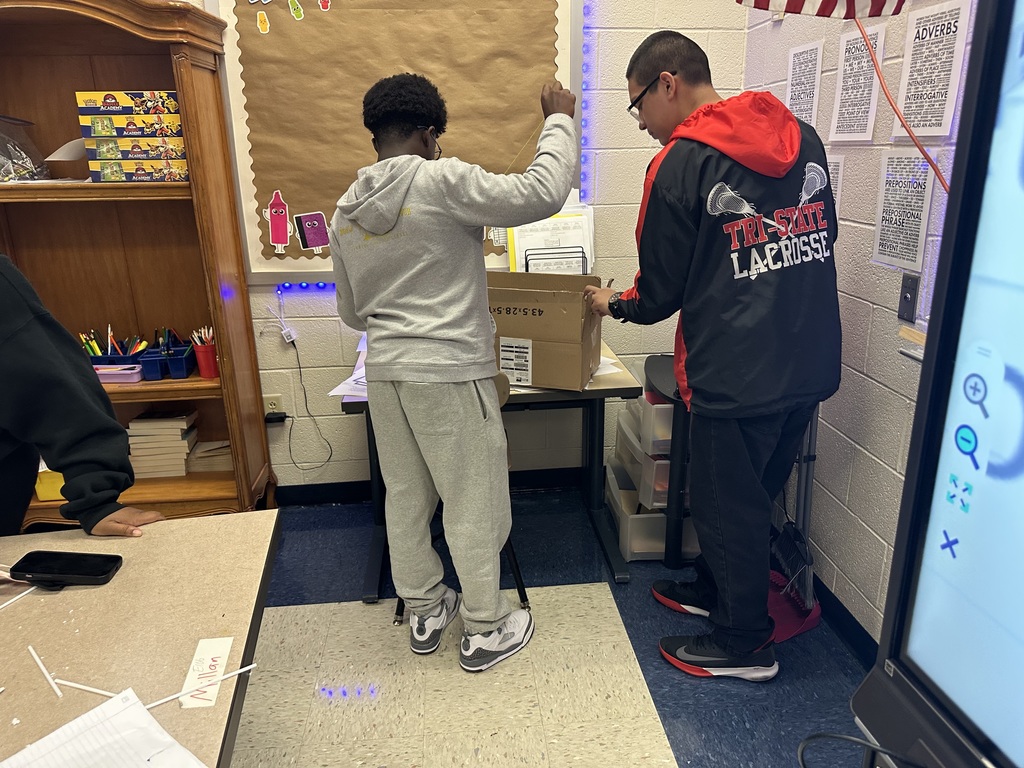 Two students are standing in a classroom, collaborating on unpacking a cardboard box on a desk. The room is filled with shelves, papers, and educational decor.