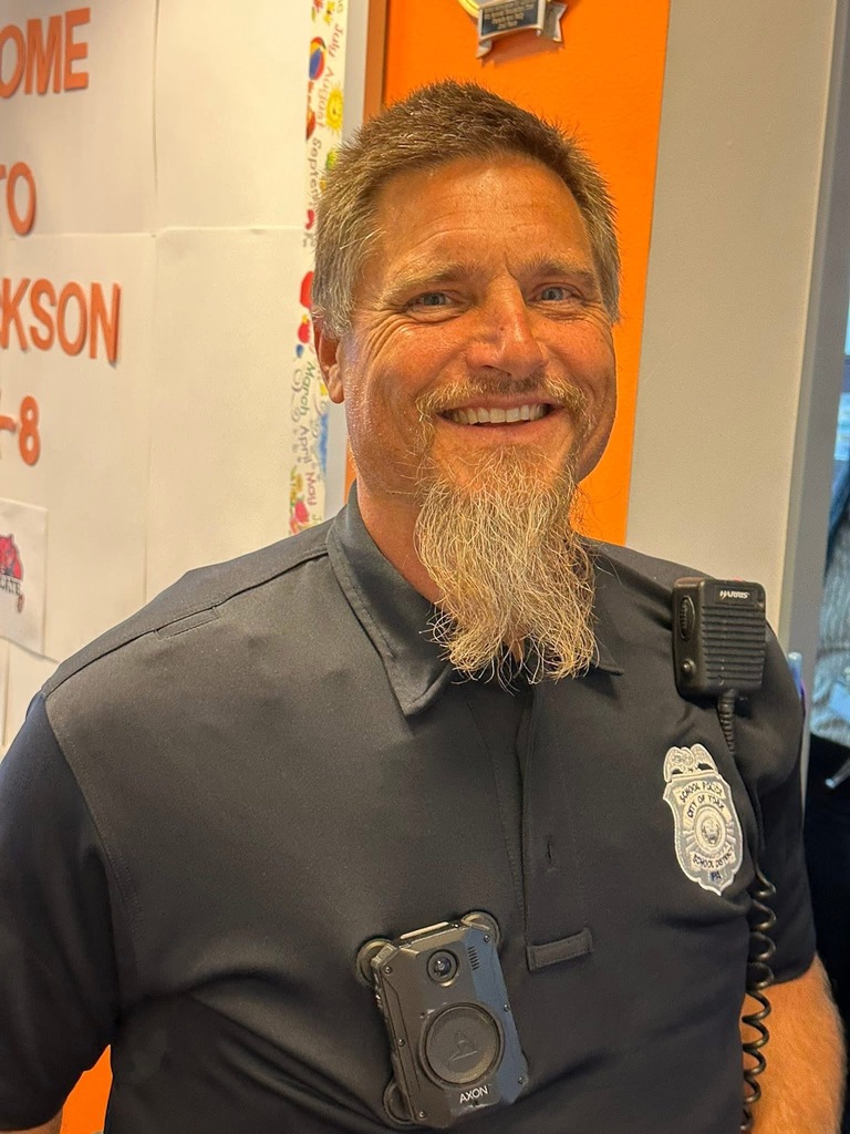 A district staff member wearing a police uniform with a badge and body camera is smiling while standing in front of a welcome sign that is in the background.