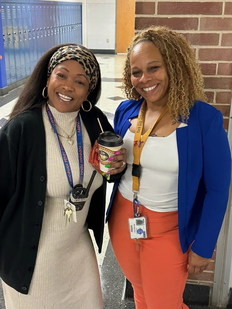Two district staff members are smiling in a school hallway. Both women are wearing lanyards.