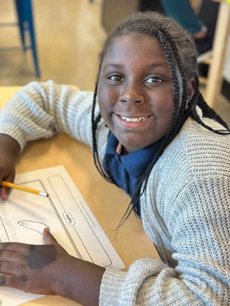 A female student is smiling while sitting at a wooden table, holding a pencil. 
