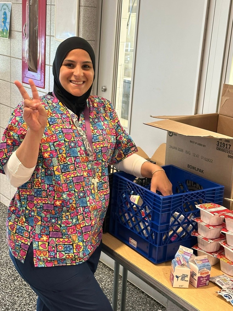 A district staff member is holding up the peace sign. She is standing by a table with a crate of milk cartons and mini containers of cereal. 