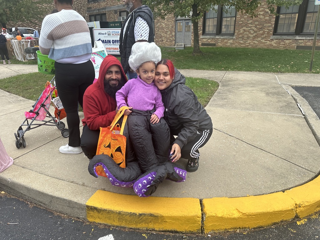A child dressed in a purple costume is standing near a yellow curb, surrounded by two adults in cozy outfits during a festive, community Halloween gathering.
