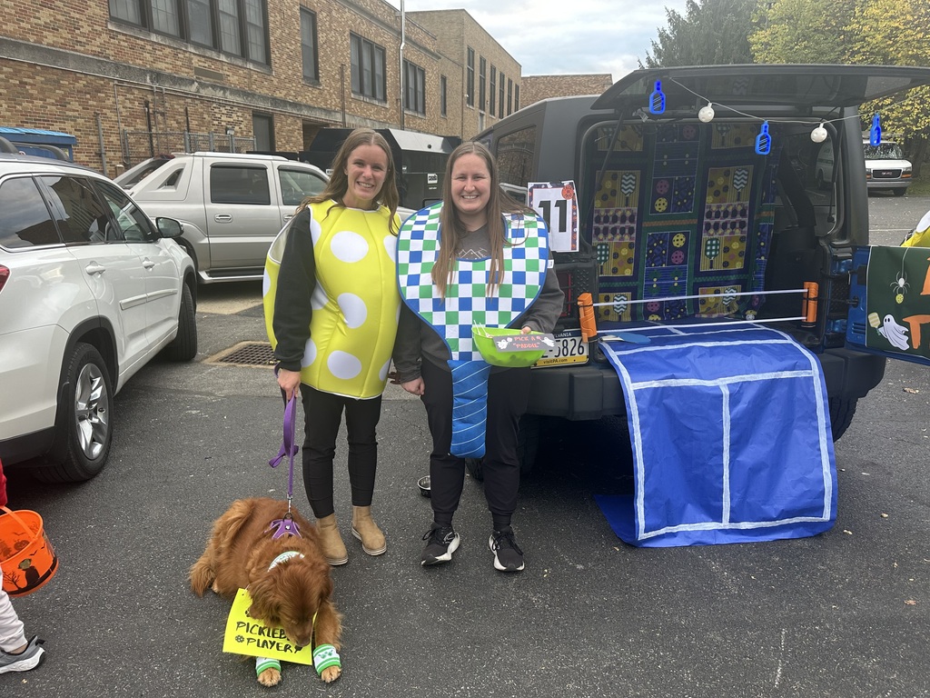 Two people in playful costumes are standing next to a decorated vehicle featuring a game setup and a dog is wearing a sign that reads "Pickler Player."