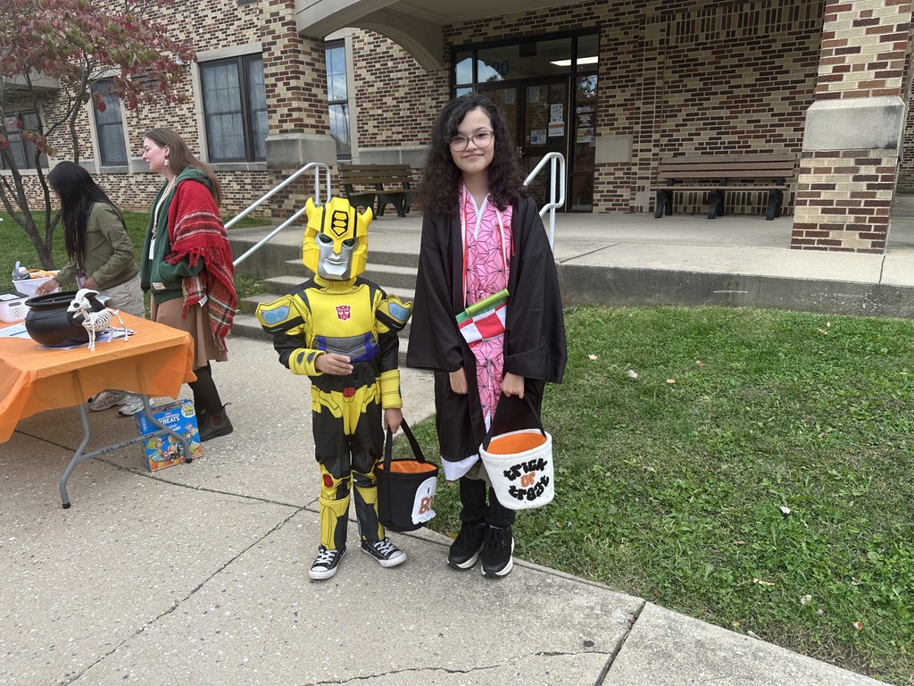 Two children in Halloween costumes are standing on a pavement. One child is dressed as a yellow robot, the other child in a brown cloak, holding treat bags. A table with Halloween decorations is nearby, and two adults are in the background.
