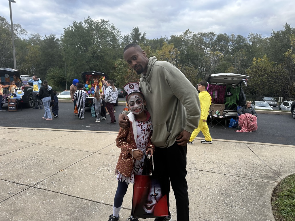 A man and a child in spooky clown makeup are posing outdoors during a Halloween trunk-or-treat event. Cars with decorations and people in costumes are in the background.