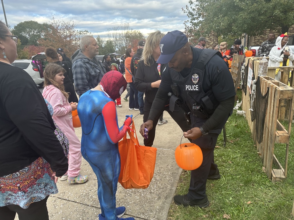 A child in a Spider-Man costume trick-or-treats is being given candy by a school police officer who is handing out candy from a pumpkin bucket during a festive, community Halloween gathering.
