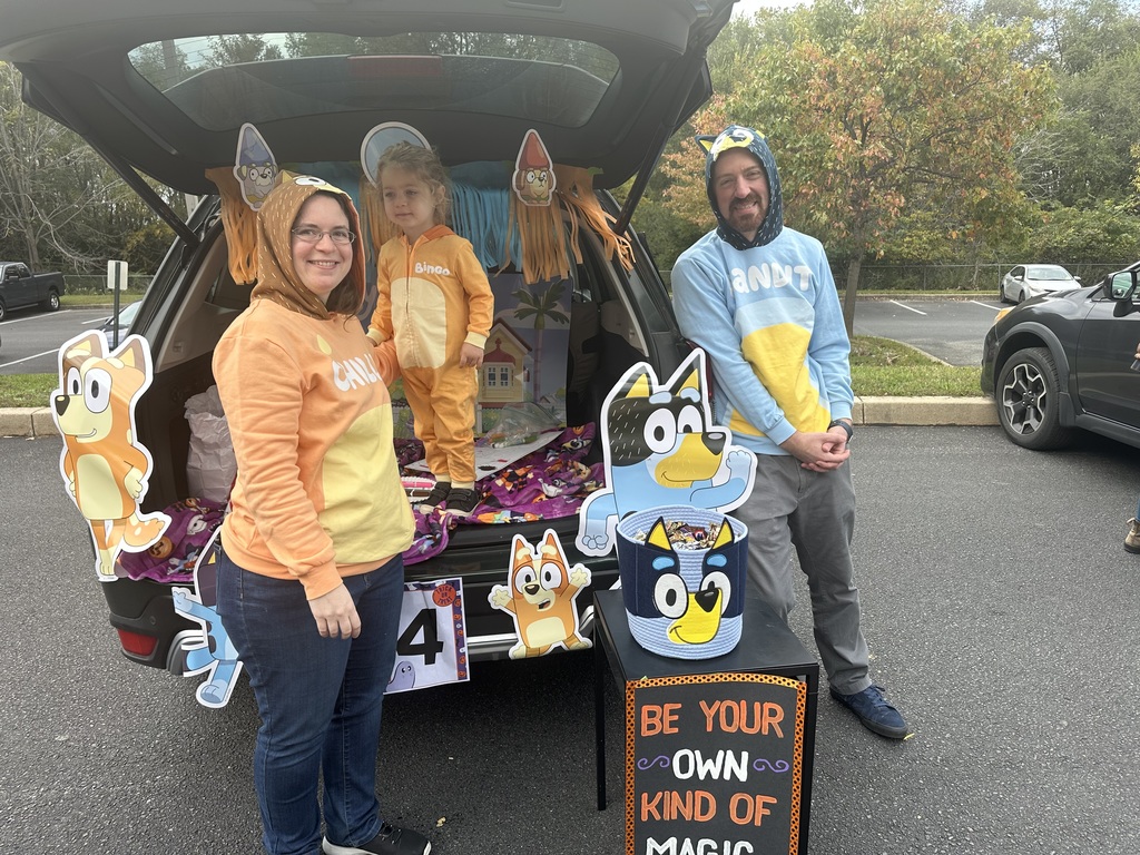 A family dressed in cartoon-themed costumes are standing around a decorated trunk at a Halloween event. They are smiling warmly, surrounded by colorful cutouts.