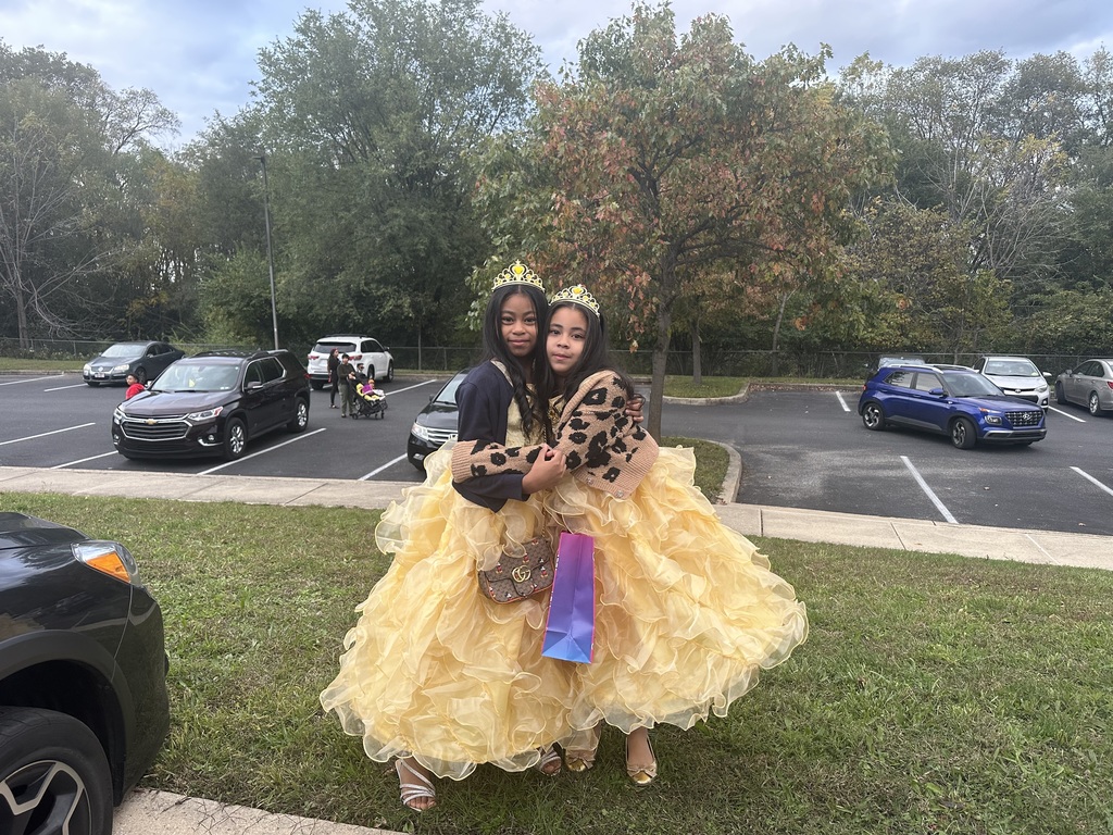 Two young girls dressed as princesses are posing together outdoors in a parking lot, wearing matching golden-yellow ball gowns with voluminous ruffled skirts and gold tiaras. One girl is wearing a tan leopard-print jacket and holding a colorful gift bag, while the other girl has a purse over her shoulder. Several cars can be seen in a parking lot in the background.