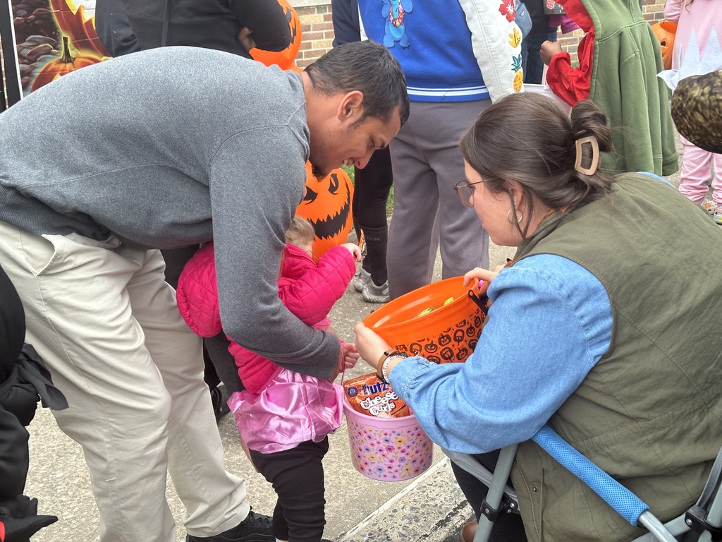 A young child in a pink coat is holding a candy bucket, helped by a man kneeling beside them. A woman is handing out treats from an orange pumpkin bucket, surrounded by Halloween decor.