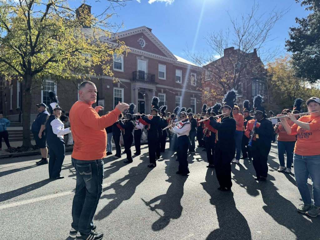 A vibrant marching band dressed in uniforms performs on a sunny street, with spectators watching and celebrating.
