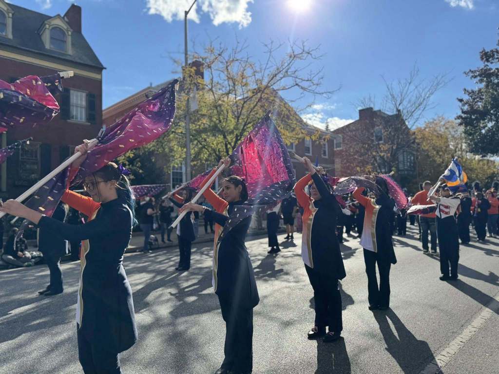 Colorful flags wave in the air as a marching band performs in a sunlit street, surrounded by autumn foliage and onlookers.